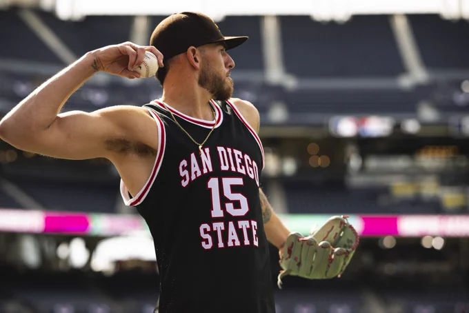 Photo of Joe Musgrove throwing a baseball. He is wearing a brown Padres hat and a black and red San Diego State basketball jersey with the number 15.