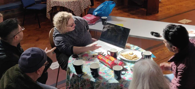 Five people sit at a round table having tea and biscuits, one of them is showing some images to the group using a laptop 
