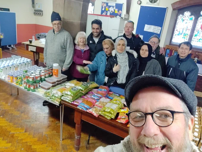 A group of men and women stand behind a table in a church hall which has a range of food on it in cans and packets on it. A man wearing a cap and glasses is in the foreground, you can see only his head.