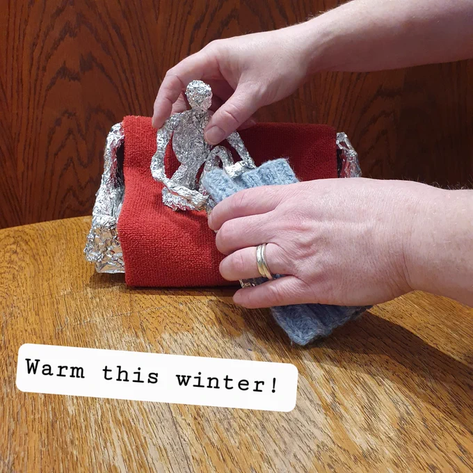 Two hands are holding a silver foil model of a man sitting on a settee with a blanket.