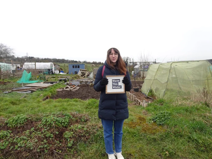 A woman is standing next to some allotments holding up a sign that says 'Warm this Winter'.