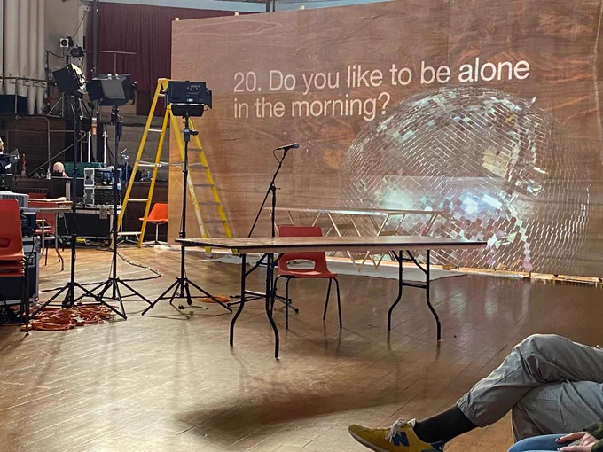 Looking out into a hall with microphones, lights and a ladder around the space. A wooden structure has a disco ball projected on it, with a question - 20. Do you like to be alone in the morning? 