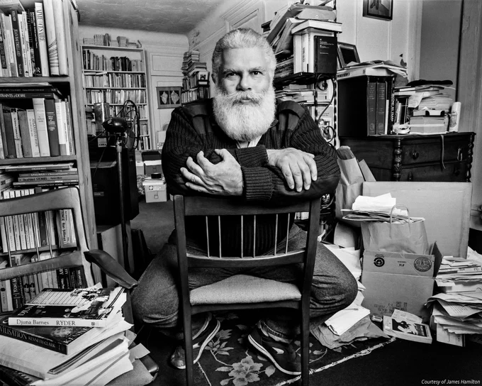 Black and white photo of Samuel “Chip” Delany sitting in a room surrounded by books on shelves and on the floor around him. He is sitting reverse on a chair with his hands folded over the chair back, staring into the camera. 