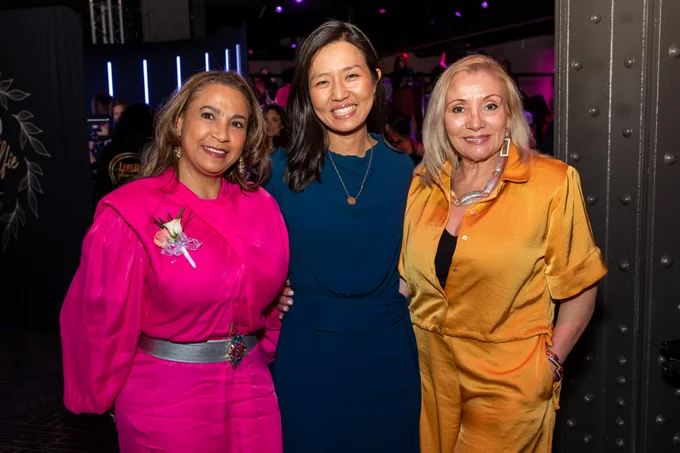 Mayor Wu, wearing a navy blue dress & gold pendant necklace, stands in-between two attendees, all smiling. The attendee on the left is wearing a bright pink dress, while the attendee on the right wears a gold jumpsuit.
