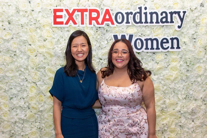 Mayor Wu stands next to an attendee in front of a wall made of white flowers. Text above them on the wall says “EXTRAOrdinary Women”, with “extra” in red and the rest of the letters in navy blue.