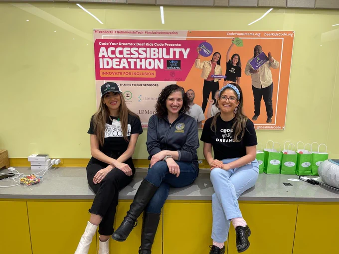 Shireen Hafeez of Deaf Kids Code, MOPD Commissioner Rachel Arfa and Brianne Caplin of Code Your Dreams are sitting on a counter posing for a photo below a sign with the words “Accessibility Ideathon”