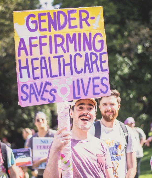 Robbie and Jake are smiling. Robbie holds a sign that reads “Gender-affirming healthcare saves lives”