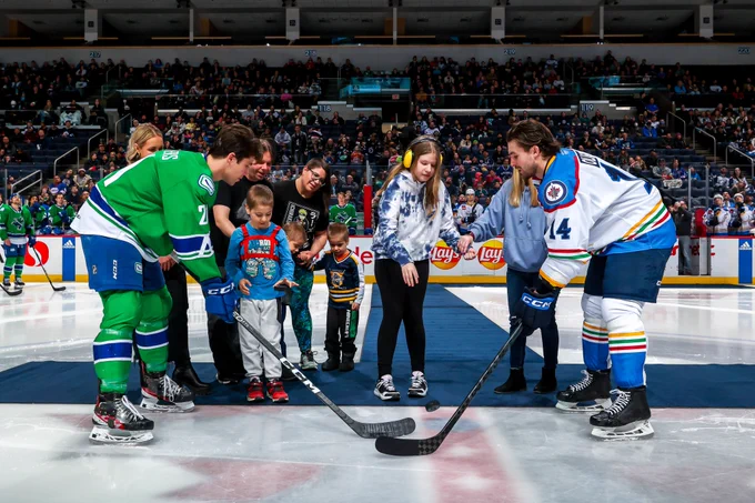 Hockey players participate in a ceremonial puck drop at the centre ice with a child and a teenager dropping pucks as their families watch.