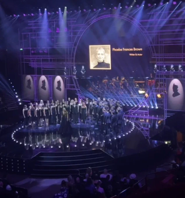 The Olivier Awards. Projected: an image of a young white woman with short hair and the words 'Phoebe Frances Brown Writer & Actor'. Below the projection are an ensemble dressed in black and standing in a semi circle on a stage.  