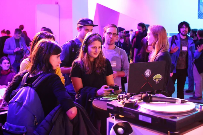 A picture taken by Julian Dasgupta on November 5, 2019 at Talk & Play #39. It shows a person playing the game Kraken Academy!! on a laptop using a gamepad while 4 other people are observing the screen. Annika Maar (one of the people behind the game) is talking with the group while looking in the other direction. In the background more people can be sen chatting.