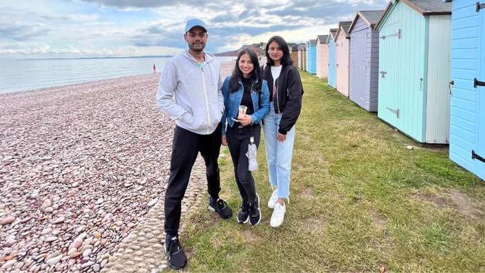 3 students stood by a pebble beach and colourful beach huts