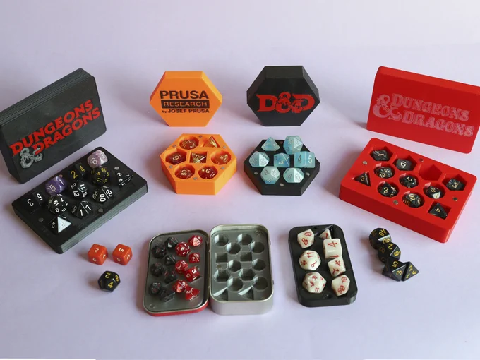 A set of different dice boxes and inserts, in two rows. All boxes are open, showing their two parts: cover serves as tray for the dice to stand on, bottom has prism-shaped holes. Upper row: A black box for 11 dice with the 5th edition D&D logo in red and silver; a hexagonal box in orange, with Prusa Research logo in black; a hexagonal black box with the short D&D 5th edition logo in red; a red box with the classic Basic red box logo in silvery white. Bottom row: three spare dice (2 d6, 1 d12); an open Altoids box with silver inserts for two sets of 7 mini dice, which are set in; one set is red with white numbers, the other black with red numbers. At the right of the Altoids box, there is another insert for an Altoids box with a full set of 7 regular-sized dice, in pearly white with ornated red numbers. At the rightmost end there are 4 black dice with yellow numbers (2 d10, 1 d8 and 1 d4).