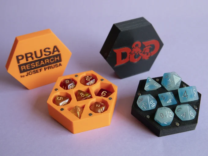 Two hexagonal boxes holding a 7-piece set of dice each. Left one is orange, with the Prusa Research logo in black, having orange glittery dice in it. Right one is black, with D&D logo in red, holding an icy blue set of dice in ready-to-pick position.