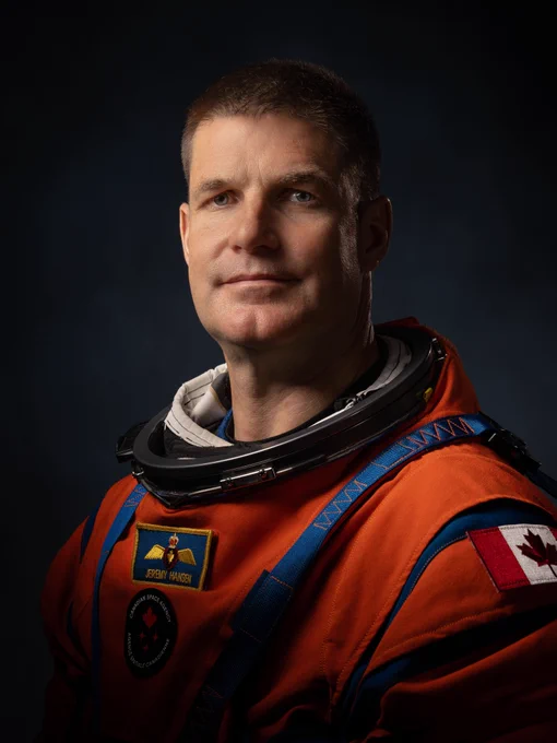 Canadian astronaut Jeremy Hansen poses for a portrait in his orange Artemis flight suit. The suit has blue trim around the neck and shoulders, with three patches: one with the Canadian flag on his left shoulder, one with his name and a pair of wings on his chest, and one bearing the logo of the Canadian Space Agency faintly visible beneath the second. The background is dark, and the photo is lit to focus on Hansen’s face, which is facing the camera with a dignified expression. Credit: NASA
