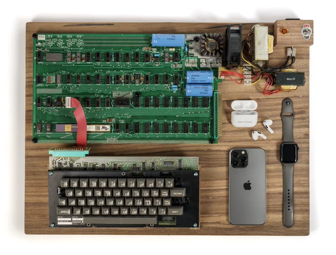 A 1976 Apple-1 Computer along with a modern iPhone, Apple Watch, and AirPods.