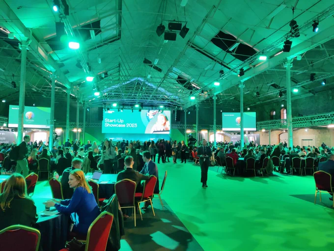 Picture of a large room within the RDS Dublin with many seated attendees surrounding circular tables. The carpet and lighting are green and large screens are situated at the front with a stage at the center.