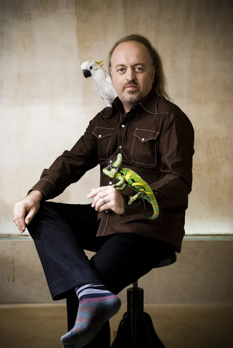 [IMAGE] A man sits on a stool and looks at the camera. He has a parrot on his shoulder and a chameleon on his other hand. He wears a brown western shirt, black trousers and stripy socks. 