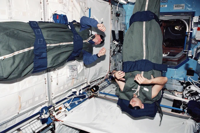 Astronauts Thomas D. Jones and Mark L. Polansky, STS-98 mission specialists, are photographed during their sleep shift aboard the International Space Station (ISS). Jones is sleeping at a 90-degree angle strapped to an interior wall on the left side of the image. Polansky floats at a 180-degree angle upside-down vertically. Both crewmembers wear sleeping masks and are in sleeping bags. The interior wall compartments of the space station module are padded white with instrument cables exposed.

Credit: NASA