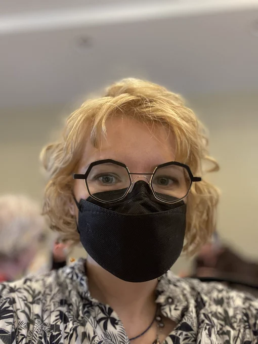Woman with short blonde wavy hair wearing black glasses and black mask