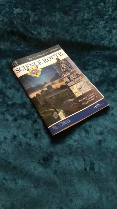 Photograph of a booklet on a green/blue background - there are photos of observatories and statues on the cover and the title is ‘city walking tour of Leiden: Science Route. Discover Leiden European City of Science’