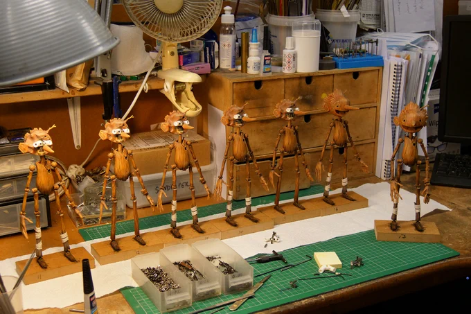 Color photo of a row of Pinocchio puppets on a green cutting mat on a workbench in various states of assembly surrounded by parts and materials.