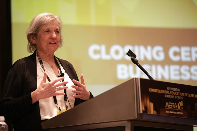 Felice Levine stands at a podium and microphone in front of a screen display the Closing Ceremony slide.