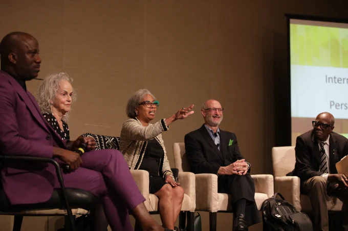 Four individuals sit on lounge chairs on a stage. One of the individuals in the middle is gesturing with her hand and speaking.