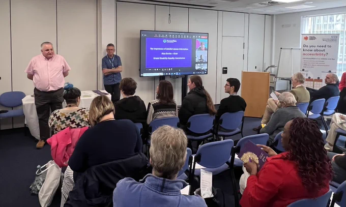 Alun Davies (Chair of Bristol Disability Equality Commission) talking to an audience at UHBW's AccessAble launch event. Alun is at the front of the room with a PowerPoint screen to the right of him. He is stood in front of three rows of people.