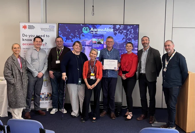 Group photo of UHBW and AccessAble colleagues with local partners receiving an AccessAble plaque. There are 9 people in a line with a PowerPoint slide and AccessAble banner stand behind them.
