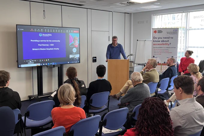 Bristol & Weston Hospitals Charity's CEO Paul Kearney talking to the audience at UHBW's AccessAble launch event. Paul is stood in front of a rostrum with a PowerPoint presentation to the left and a banner stand to the right.