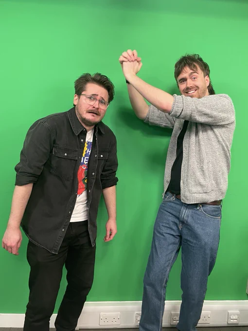 Tom B and Luke stand in the Yogscast livestream studio in front of a green wall. Luke it pretending to bonk Tom on the head. Luke smiles with Tom looks dismayed.