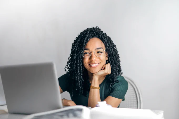 Black female student sitting at table with laptop, posed with hand on chin and smiling.