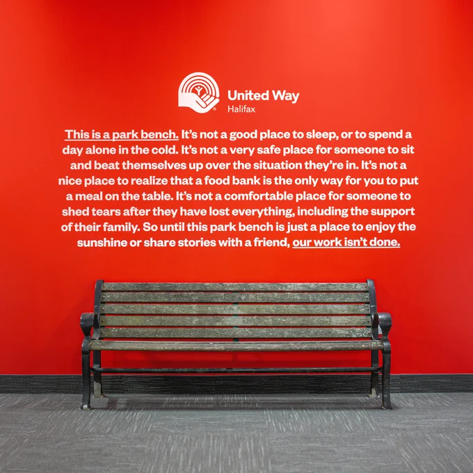 A bright red office wall with a weathered park bench sitting in front of it. Above the bench, white text reads: "This is a park bench. It's not a good place to sleep, or to spend a day alone in the cold. It's not a very safe place for someone to sit and beat themselves up about the situation they're in.  It's not a nice place to realize that a food bank is the only way for you to put a meal on the table. It's not a comfortable place for someone to shed tears after they have lost everything, including the support of their family. So until this park bench is just a place to enjoy the sunshine or share stories with a friend, our work here isn't done." Above that text is a white United Way Halifax logo.