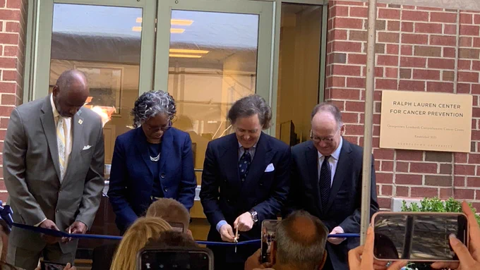 Four people stand in front of a brick building. They all hold gold scissors, and are using them to cut a blue ribbon. On the building, there is a sign reading "Ralph Lauren Center for Cancer Prevention".