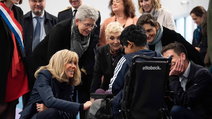 Photographie sur laquelle sont visibles Geneviève Darrieussecq, Brigitte Macron et Amélie Oudéa-Castéra. Les trois femmes souriantes s'adressent à une jeune personne en fauteuil roulant, face à elles. En arrière-plan, d'autres personnes sont présentes dont l'une d'entre elles porte une écharpe tricolore.