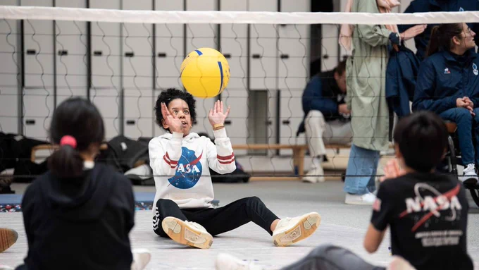 Photographie d'enfants en situation de handicap jouant au volley-ball assis. L'un des trois s'apprête à renvoyer le ballon de l'autre côté du filet. 

En arrière-plan, d'autres personnes sont présentes dont une assise dans un fauteuil roulant.