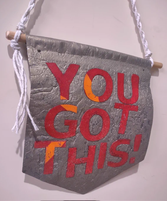 Small gold plastic banner with white string and wooden dowel, with red and orange plastic letters reading "You got this!"