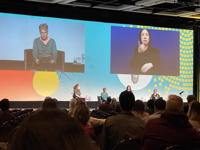 Dr. Tal Jarus sharing her experiences and research findings to discuss how people with disabilities experience medicine. Panel on a stage, big screen of Tal Jarus, woman in green blouse and interpreter in black.