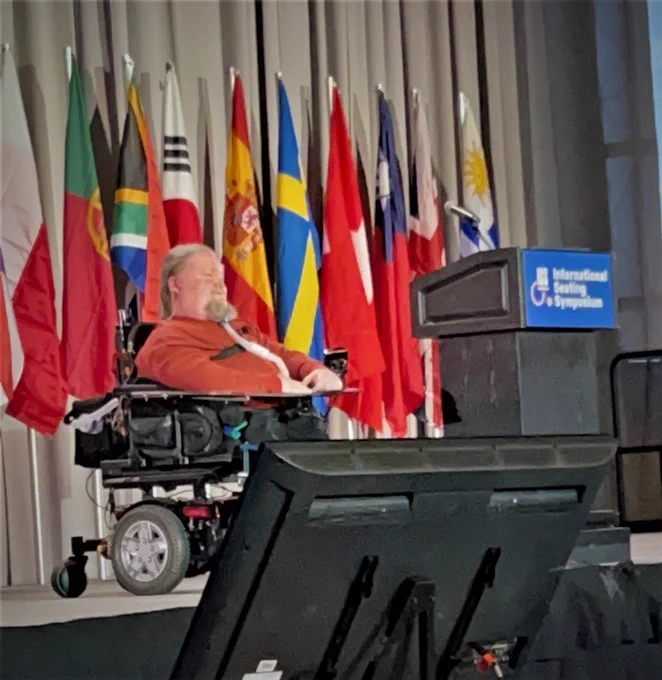 Steve wearing a red shirt and Sitting in a black wheelchair on stage with a giant podium, floor monitor in front of him, and flags from around the world behind him.