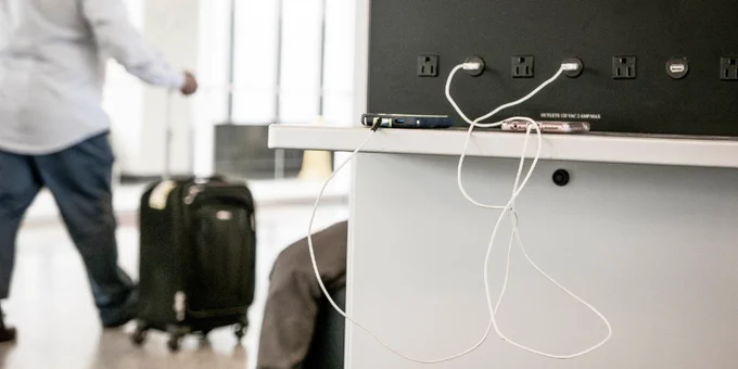 Photo shows person walking with a rolling luggage item and two phones plugged into a USB charging wall