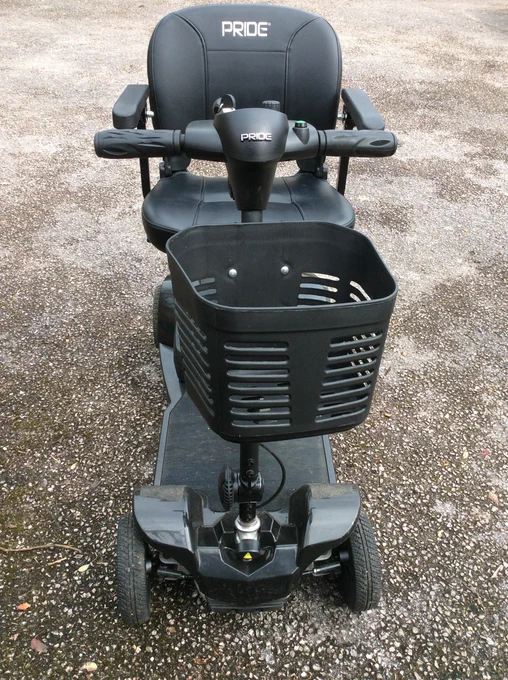 Black mobility scooter facing forward with a basket attached in front. The word Pride is stamped in front of the controls and on the backrest. 