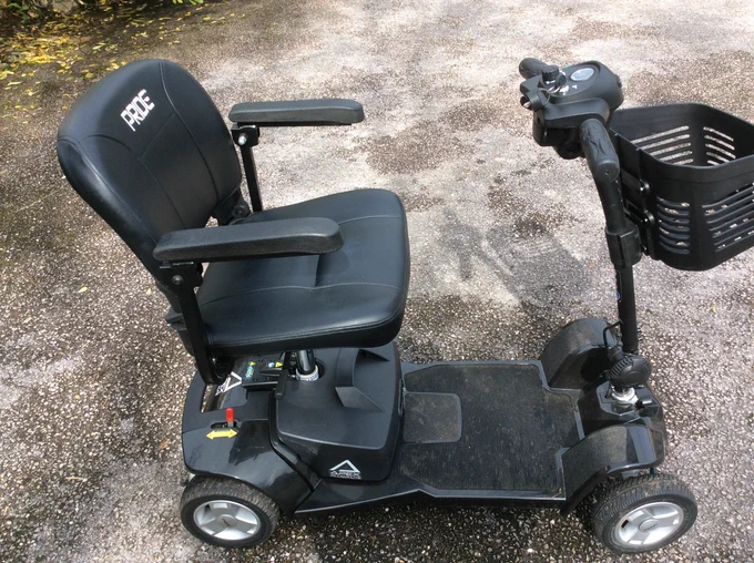 Black mobility scooter facing sideways with the wheels, seat, backrest, arms, handle bars, and basket are visible. The word Pride can be seen stamped on the backrest. 