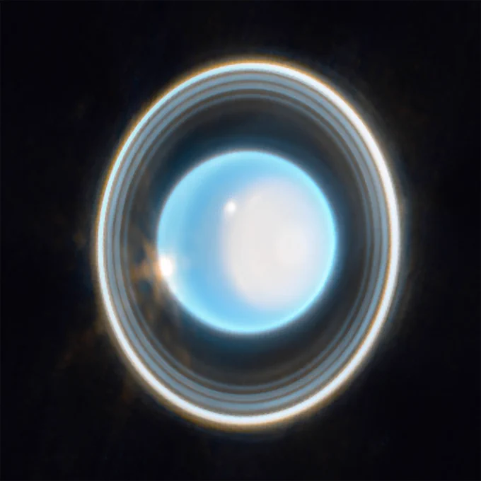 The planet Uranus on a black background. The planet appears light blue with a large, white patch on the right side. On the edge of that patch at the upper left is a bright white spot. Another white spot is located on the left side of the planet at the 9 o’clock position. Around the planet is a system of nested rings. The outermost ring is the brightest while the innermost ring is the faintest. Unlike Saturn’s horizontal rings, the rings of Uranus are vertical and so they appear to surround the planet.