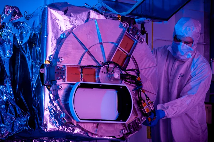 Bathed in purple light, a technician dressed in white gown, mask, cap, and gloves inspects an instrument with shiny silver color.