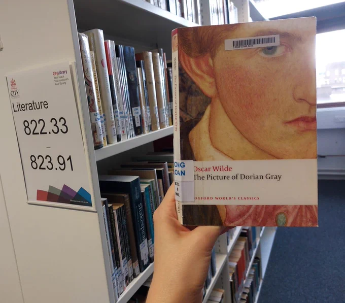 A hand is holding a print copy of The Picture of Dorian Gray. In the background, library shelves can be seen, with a shelfmark sign "Literature 822.33 - 823.91" on the nearest shelf