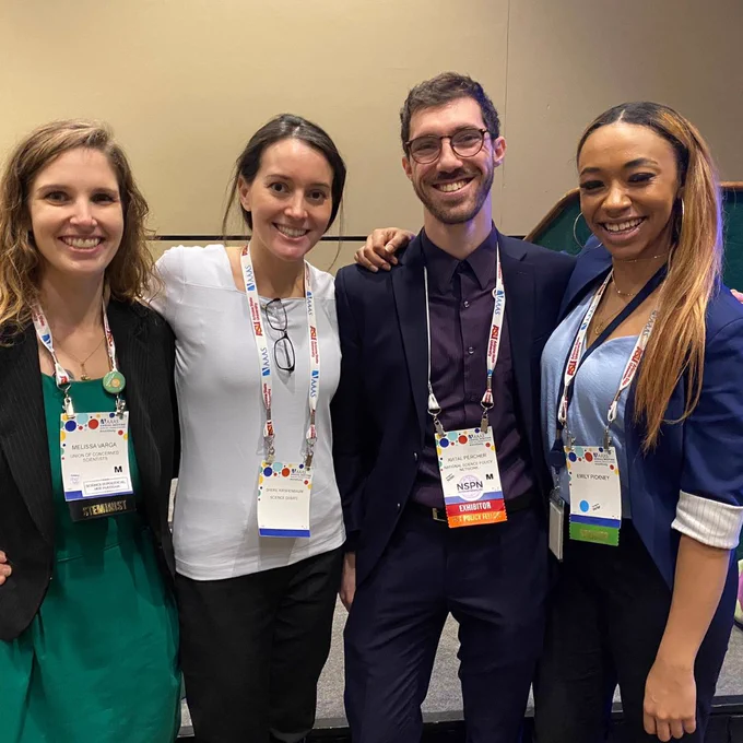 Photo of four people dressed in conference attire with their arms around one another as they pose in a line.