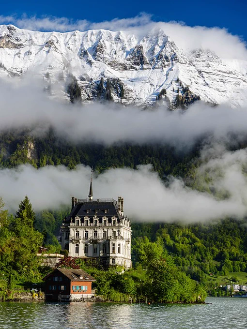 A photo of houses next to a lake at the bottom of steep mountains that are covered in snow