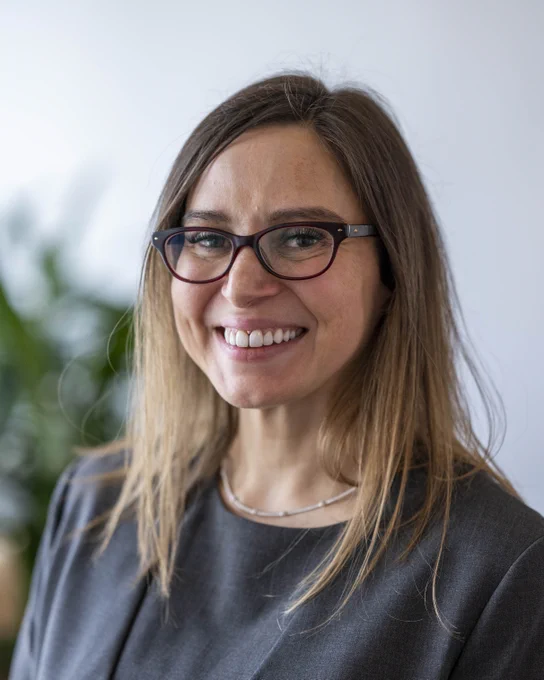 Portrait of Makenzie Lystrup, smiling at the camera. She has medium brown hair, lighter closer to the ends. She is wearing brownish, plastic-rimmed glasses, and a gray shirt and blazer. She has on a white necklace. The background is mostly white, with some out of focus greenery visible behind her right shoulder.