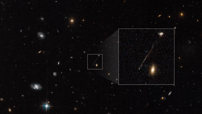 A Hubble image of a black, deep-space field is speckled with white, yellow and reddish galaxies. At the lower left corner, one, lone star with four, prominent spikes appears. In the center of the image is a small, white-bordered, boxed area that contains one, long, thin, diagonal streak of whitish-blue stars. The diagonal line looks like a scratch on the photograph. It is positioned from near the bottom, left corner to the top right corner of the box. Two galaxies also reside within the box. One irregular-looking galaxy, in the top right corner, appears to be connected to the line. Another elliptical-looking galaxy appears in the bottom, right corner of the box. To the right of the small box is a larger white-bordered box that contains a magnified view of the contents of the smaller box. In this magnified view, a white knot appears prominently at the bottom tip of the stream of stars.