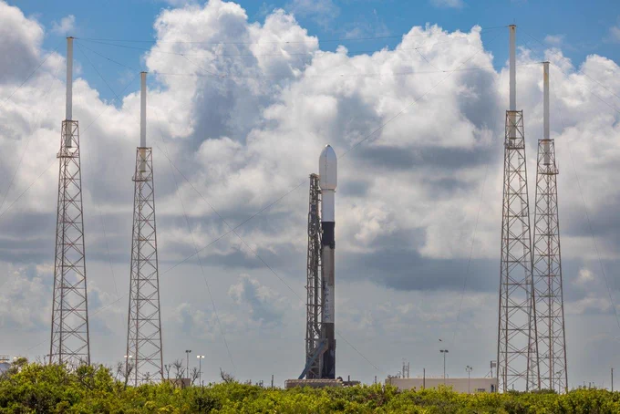 Surrounded by four towers, a launch is ready. Big white clouds billow in the sky above green foliage. 